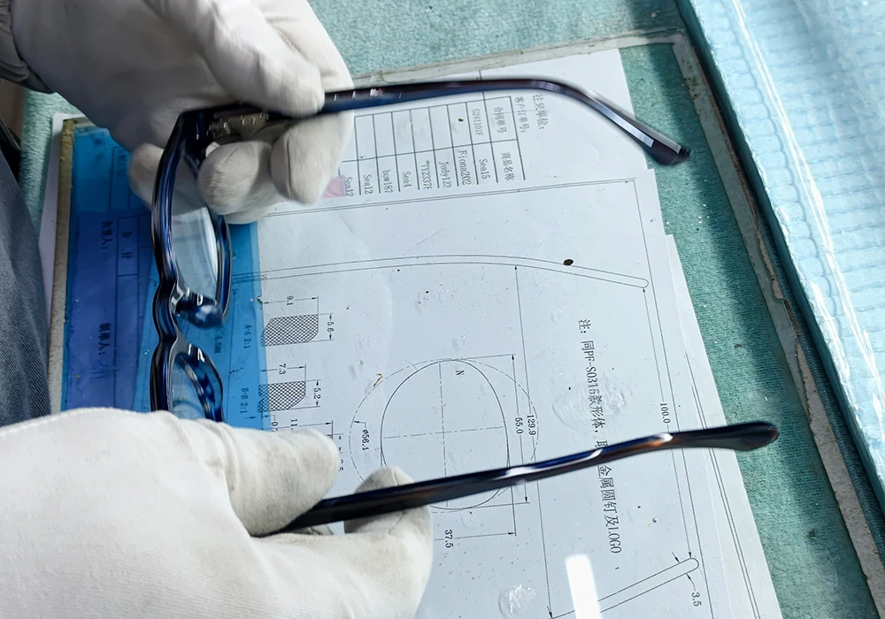 Gloved hands checking an acetate optical frame against a dimensional spec drawing during quality control